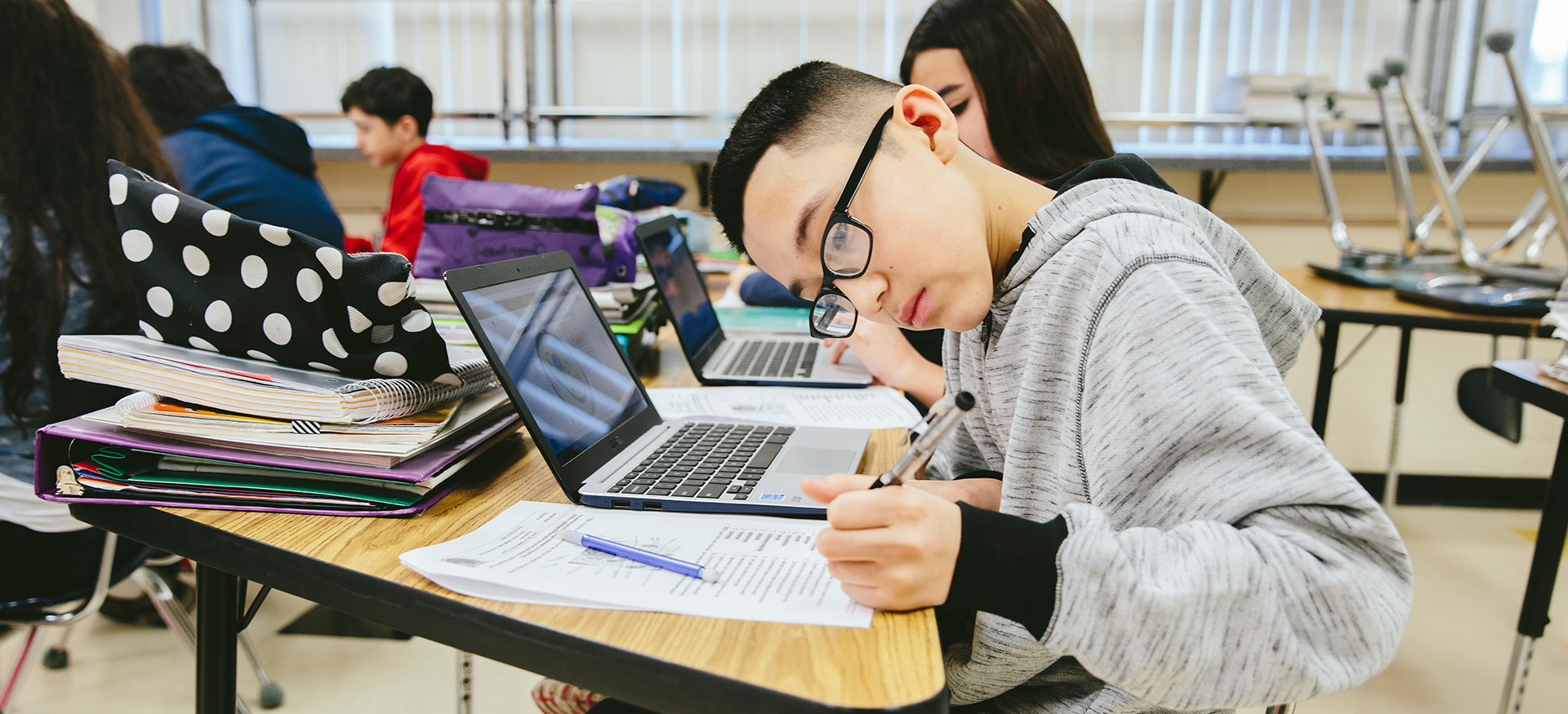 学生们用笔记本电脑在课桌上独立学习。一个学生正俯身在纸上写字。