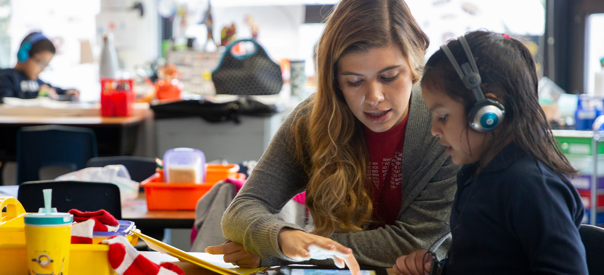 一位老师正在帮助一位戴着耳机在平板电脑上学习的年轻学生。